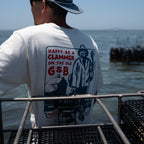 A man seen from behind wearing a white t-shirt with a large red and blue graphic print on the back that reads 'HAPPY AS A CLAMMER ON THE OLE GSB' with an illustration of a clammer, set against an outdoor water background.